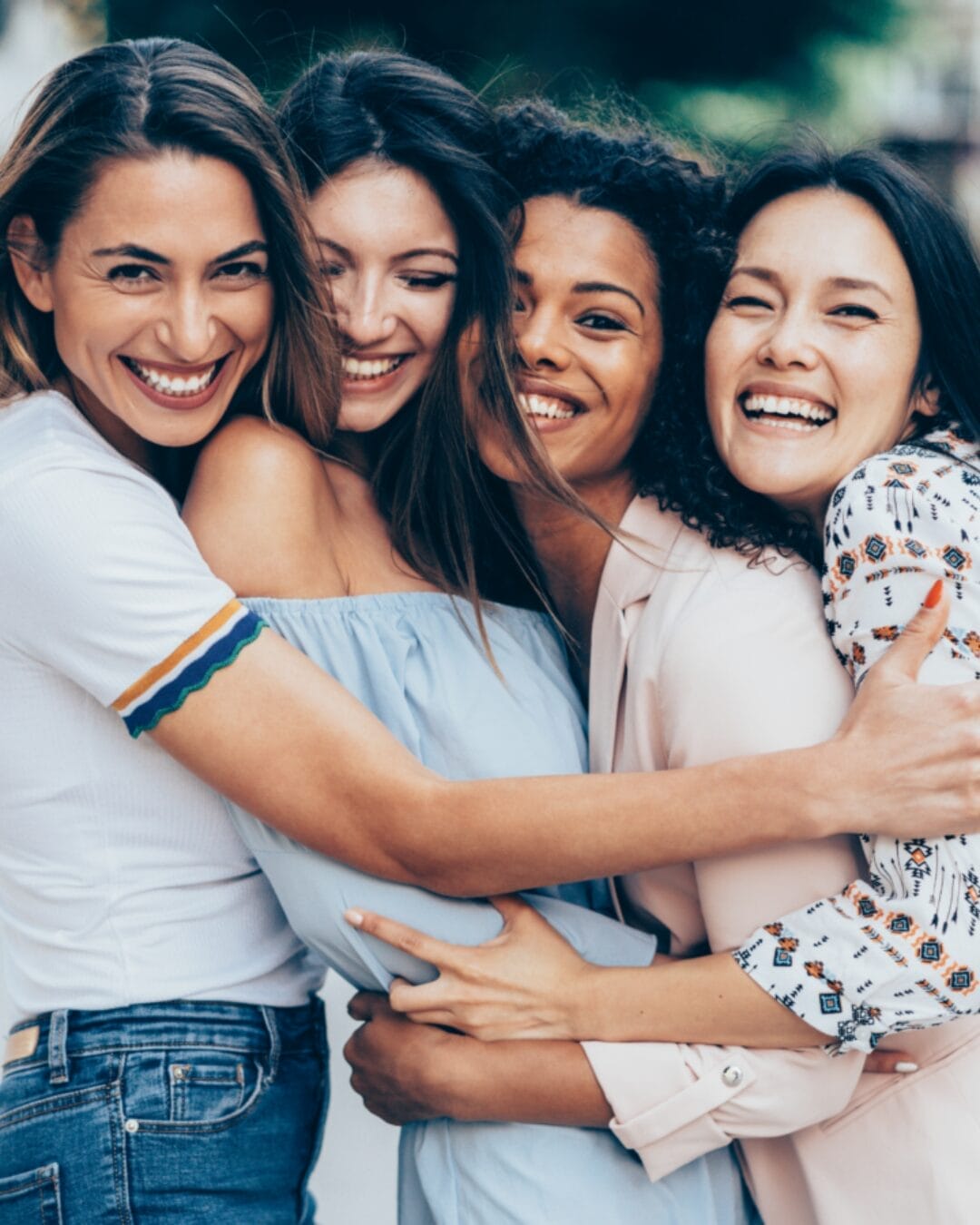 Four women embrace each other in a joyful group hug, laughing and smiling. They appear happy and connected, symbolizing friendship and empowerment.