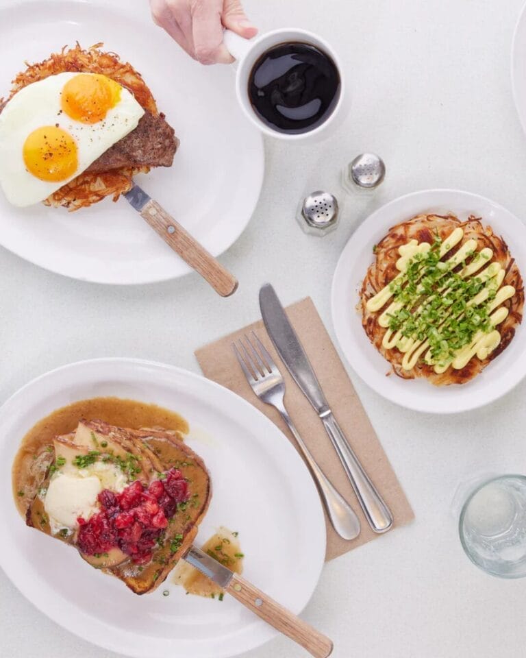 A top-down view of a breakfast spread on a white table, featuring various plated dishes. The meals include eggs with hash browns and steak, a dish with toast, gravy, and cranberry sauce, a loaded hash brown topped with sauce and chives, a plate with a sausage, bacon, eggs, and toast, and a dish with meatloaf, gravy, mushrooms, pickles, and a biscuit. A hand is seen holding a cup of black coffee, alongside silverware, a napkin, and salt and pepper shakers.