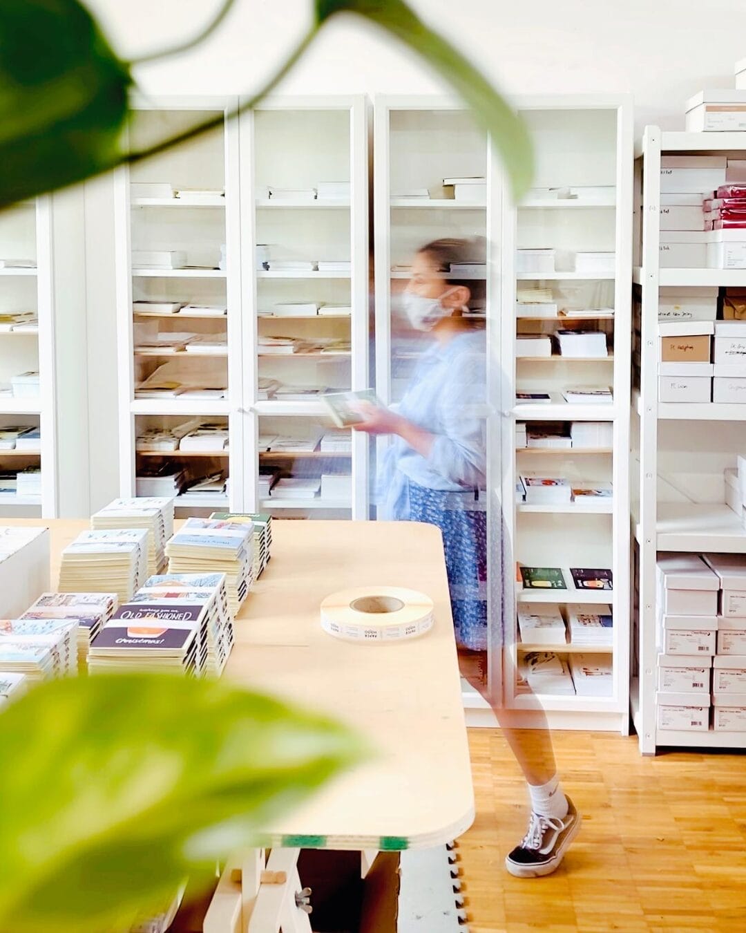 A blurred motion shot of a person wearing a face mask and casual clothing, walking through a stationery shop or warehouse. Shelves filled with neatly stacked paper goods and boxes line the background, while a wooden table in the foreground holds stacks of greeting cards and a roll of labels. A green plant leaf partially frames the image.