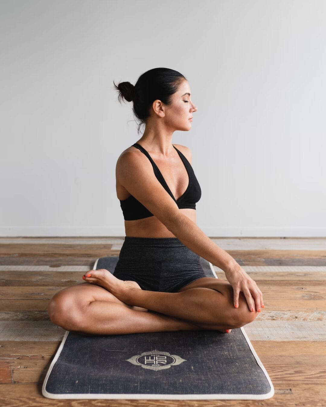 Woman in a black sports bra and shorts sitting on a yoga mat in a seated spinal twist pose. She has her eyes closed, practicing mindfulness in a minimalist space.