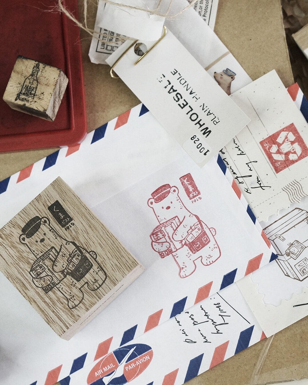 A collection of vintage-style stationery items, including an airmail envelope, stamps, and rubber stamps. A wooden rubber stamp with an illustration of a bear wearing a cap and carrying packages is prominently featured, along with a red ink imprint of the same design on the envelope.