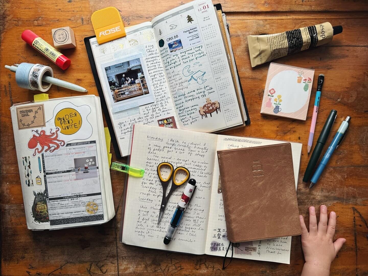 A wooden desk with an assortment of open journals, stationery, and writing tools. The journals contain handwritten notes, stickers, and a Polaroid photo. Various pens, scissors, a glue stick, washi tape, and a leather-bound notebook are scattered around. A small child's hand rests on the desk near the notebook.