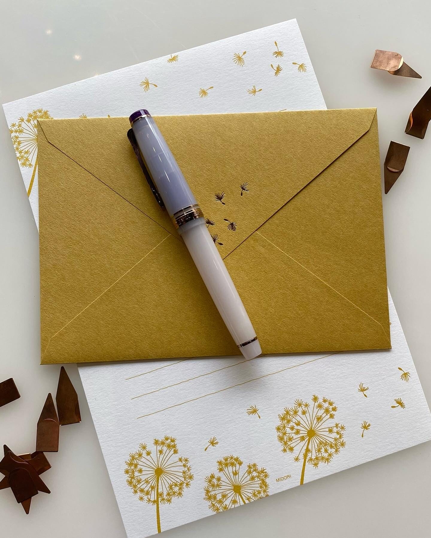 A gold envelope and matching stationery featuring delicate gold dandelion illustrations rest on a white surface. A gradient fountain pen with a purple cap lies on top, surrounded by scattered copper-colored metal clips.