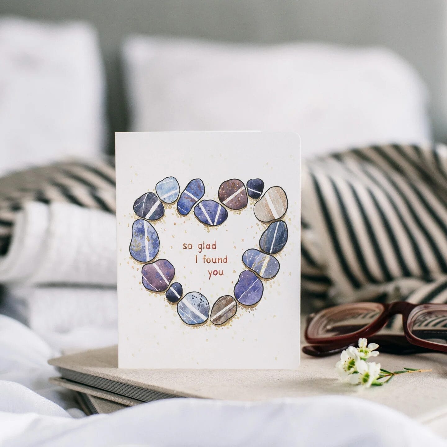 A greeting card featuring an illustration of blue and brown stones arranged in a heart shape with the message 'so glad I found you' in the center. The card is propped up on a beige book, with striped fabric, glasses, and a small white flower in the background."