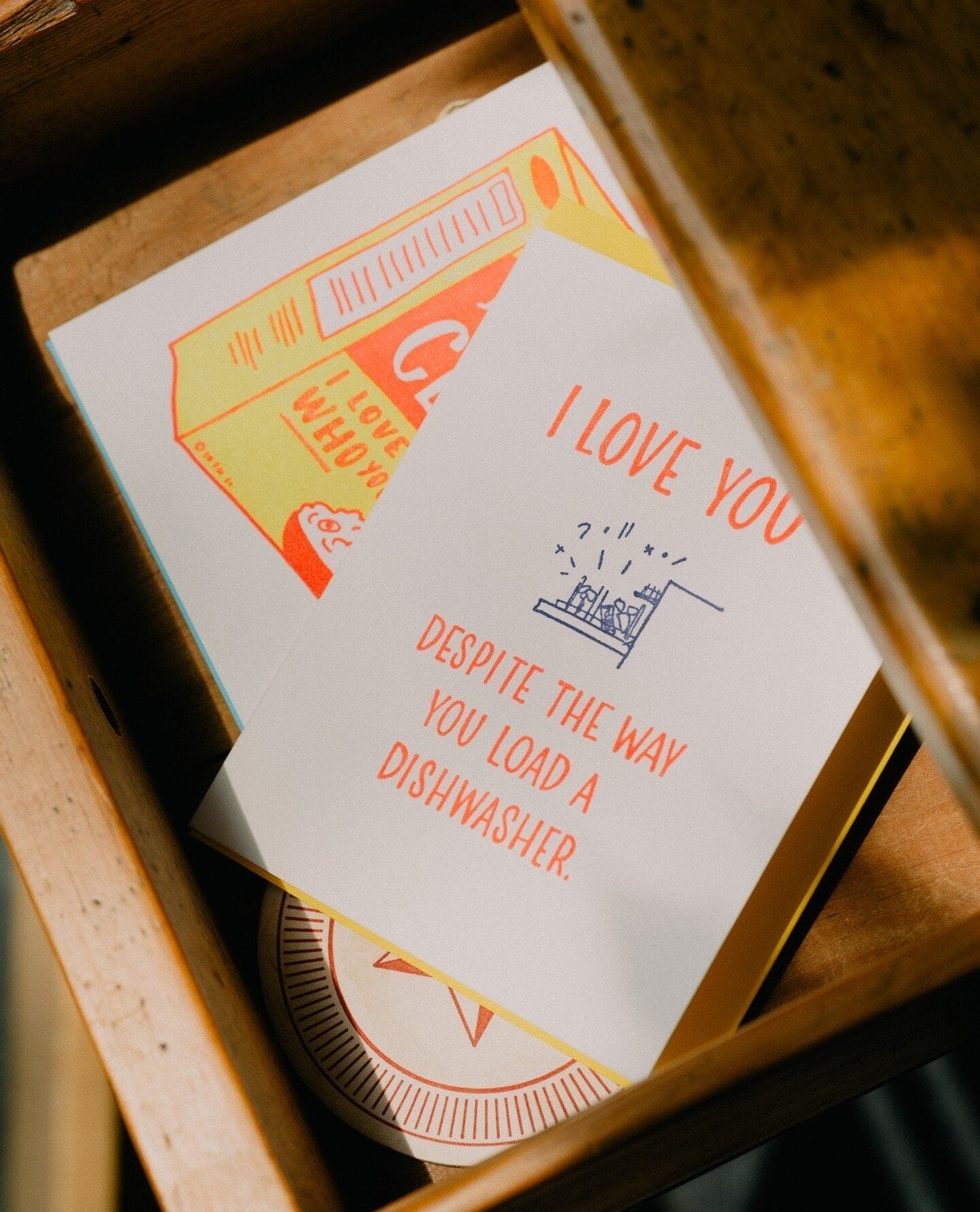 A close-up of a greeting card inside a wooden drawer. The card has bold orange and blue text that reads, 'I LOVE YOU DESPITE THE WAY YOU LOAD A DISHWASHER,' with a small illustration of a poorly loaded dishwasher. Other colorful cards with playful designs are partially visible underneath.