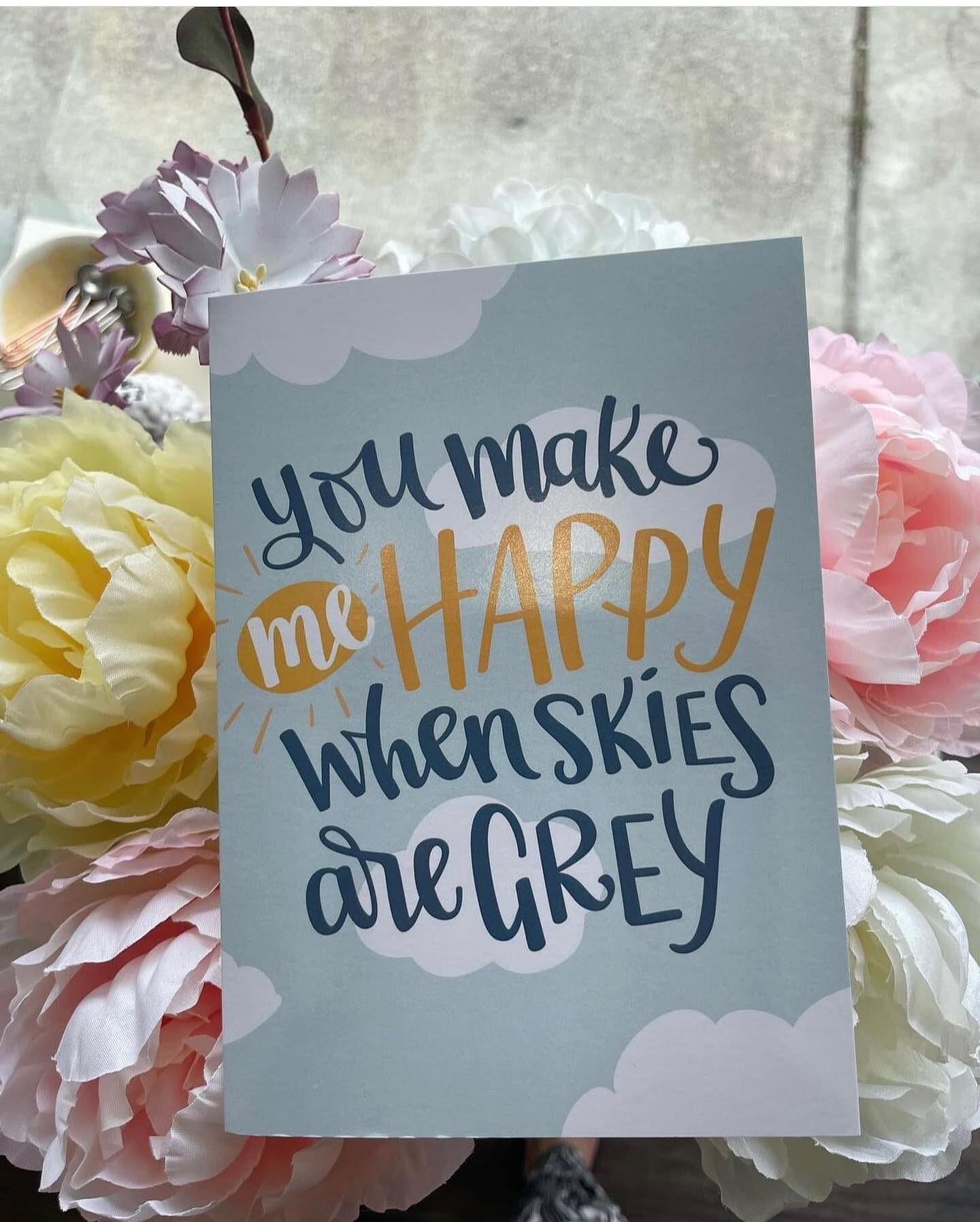 A greeting card with a light blue background and white cloud illustrations, featuring the message 'You make me HAPPY when skies are GREY' in a mix of dark blue and golden yellow lettering. The card is placed among pastel-colored artificial flowers in shades of yellow, pink, and white.