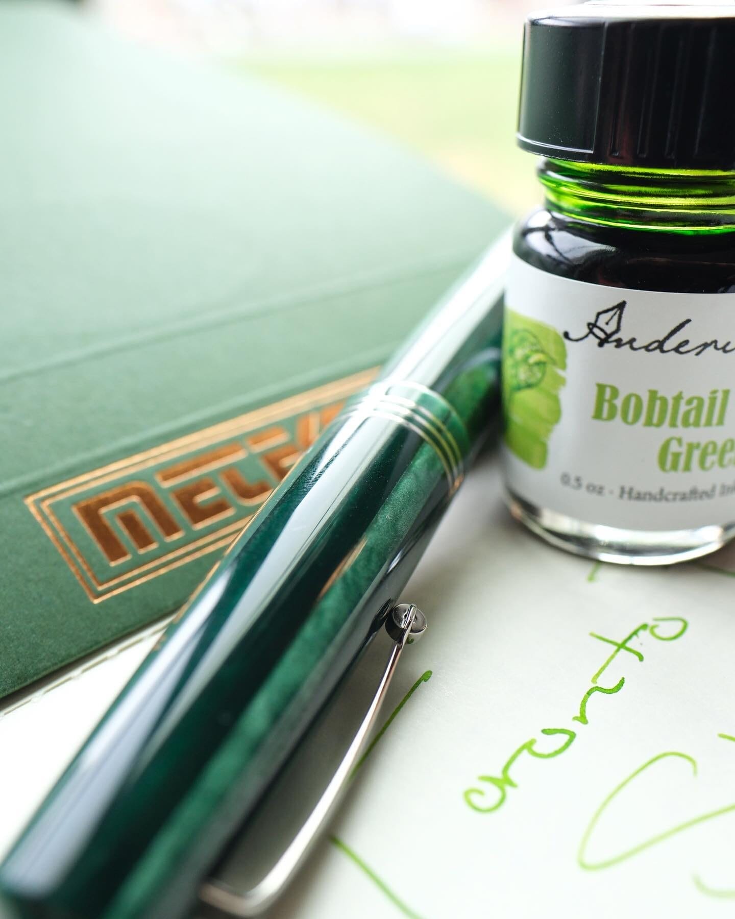 A close-up of a green fountain pen with gold accents resting on paper with green ink writing, alongside a bottle of 'Bobtail Green' ink. In the background, a green notebook with gold detailing is partially visible.
