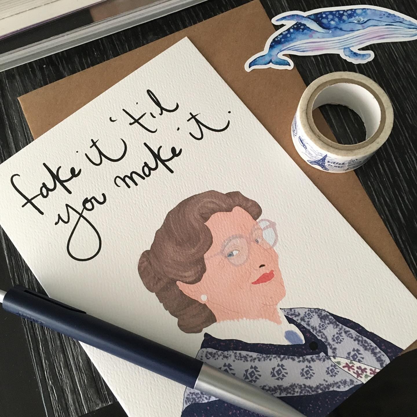 A greeting card with an illustration of a woman wearing glasses and a patterned sweater, featuring the handwritten phrase 'fake it 'til you make it.' The card is placed on a brown envelope, accompanied by a navy blue pen, decorative washi tape, and a whale sticker on a dark wooden surface.