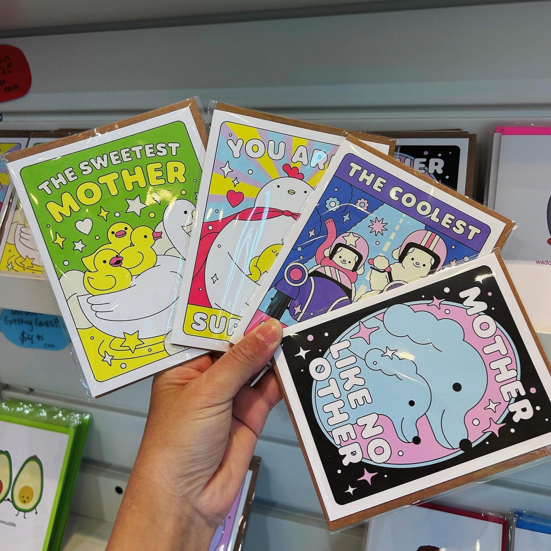 A hand holding four colorful Mother's Day greeting cards with playful cartoon illustrations of animals, each featuring a unique message such as 'The Sweetest Mother' with ducks and 'Mother Like No Other' with elephants. The background shows a display of more greeting cards on store shelves.