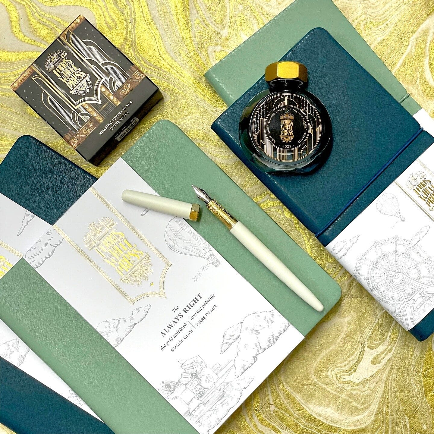 A flat lay of luxury stationery items, including green and blue notebooks, a cream-colored fountain pen with gold accents, and a black ink bottle with an ornate gold label. The background features a gold marbled surface, complementing the elegant packaging and design elements.