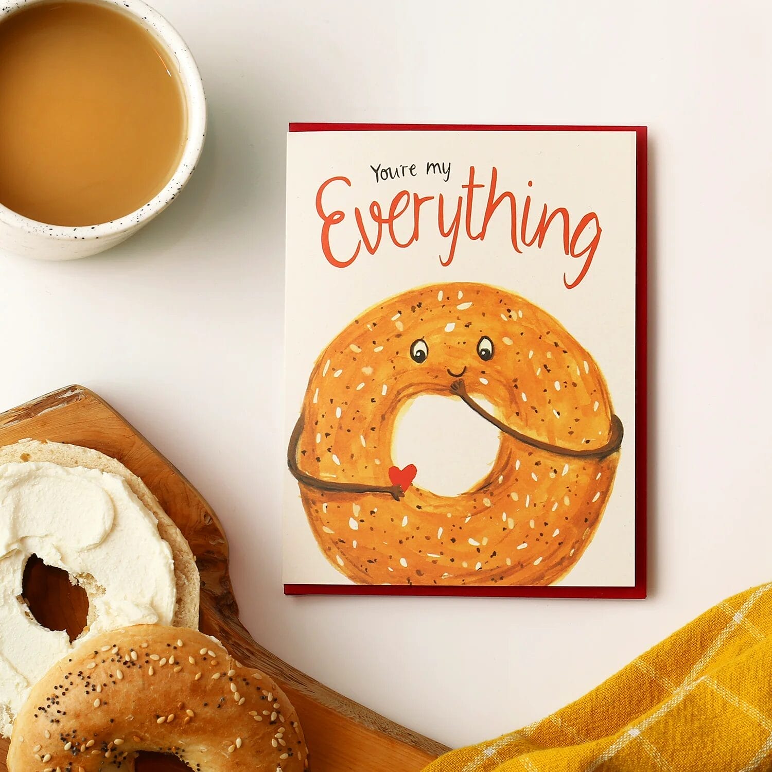 A greeting card featuring a smiling everything bagel with arms hugging itself and holding a small red heart. The card reads "You're my Everything" in playful red script. It is placed on a white surface next to a cup of tea and a wooden board with a bagel and cream cheese.