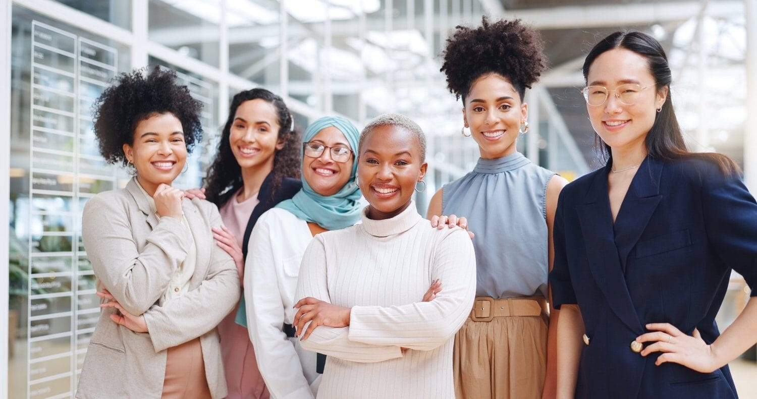 A group of six diverse professional women standing together in a modern office setting, smiling with confidence. They are dressed in business and business-casual attire, representing inclusivity and leadership.