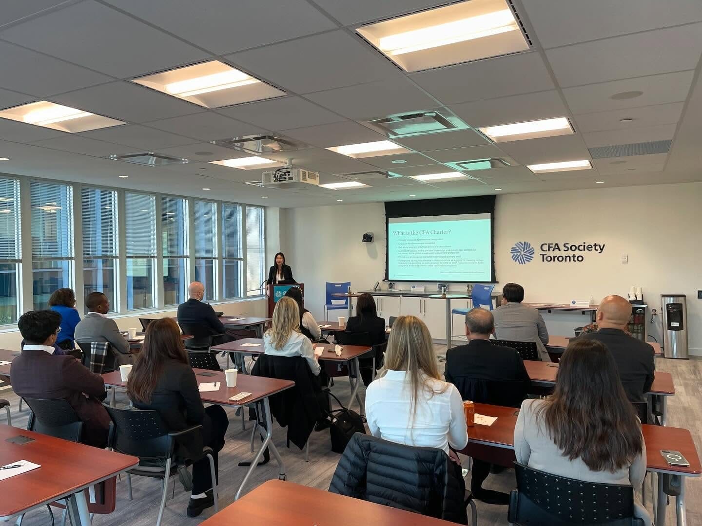 A professional seminar at CFA Society Toronto, where a woman is presenting at a podium to an audience seated at tables. The attendees are engaged, taking notes, and listening to the speaker, with a presentation slide displayed on a screen.