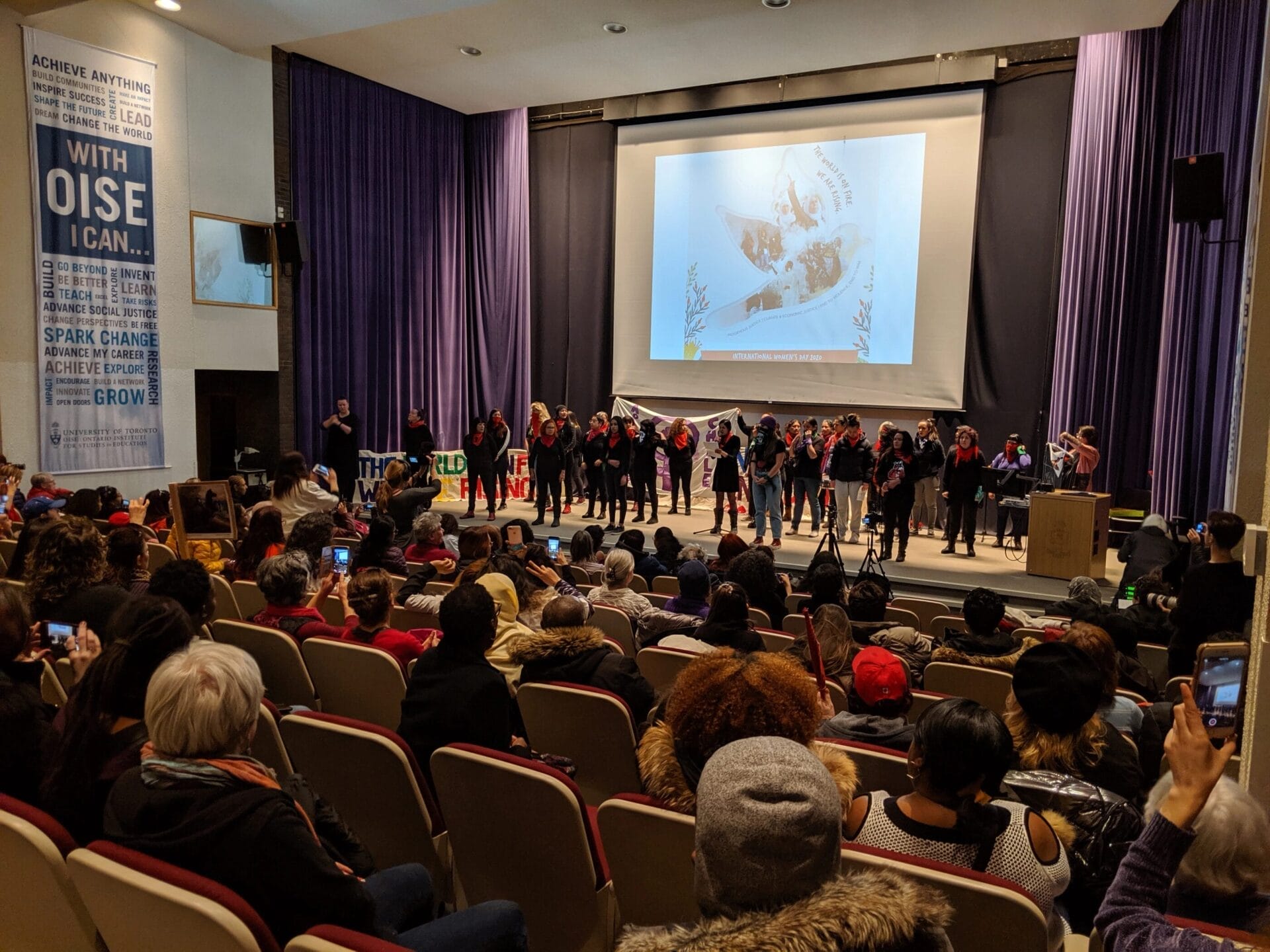 A lively event in an auditorium with a diverse audience watching a performance on stage. A group of people dressed in black and red stand in front of a large screen displaying artwork, while attendees capture the moment on their phones.
