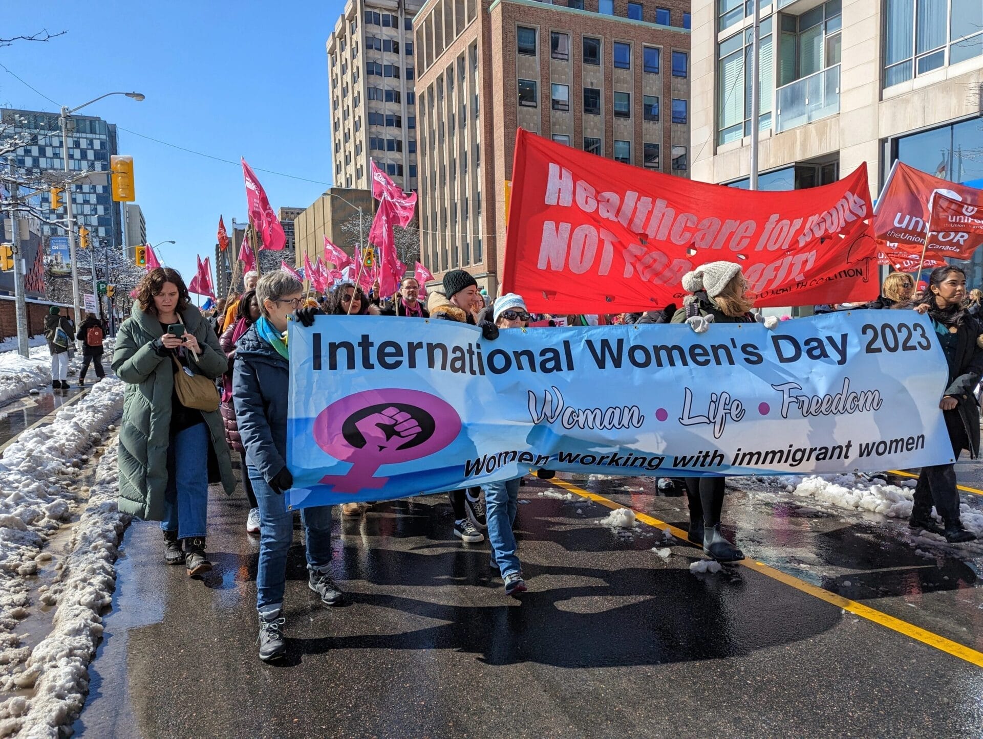 A group of people marching in a rally for International Women's Day 2023, holding a large blue banner that reads "Woman • Life • Freedom" and "Women working with immigrant women." Protesters carry pink and red flags, with a red banner in the background demanding healthcare for workers, as they walk through a snowy city street.