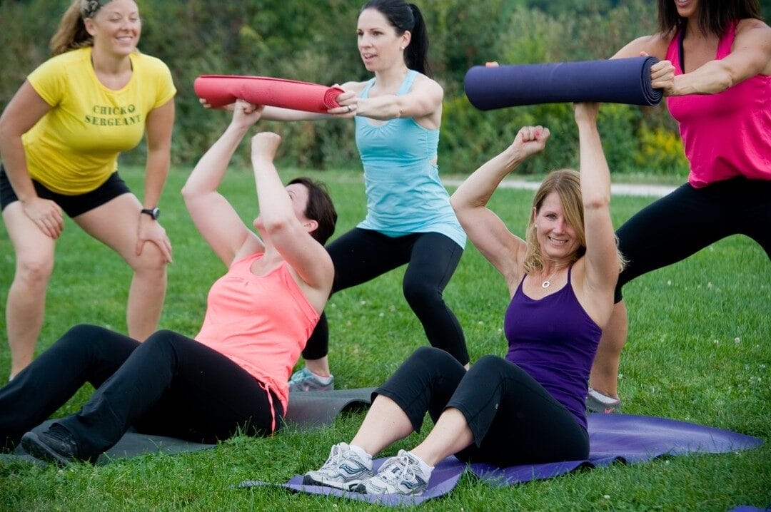Group of women exercising outdoors on a grassy field during a boot camp session. Some are seated on yoga mats performing sit-ups with rolled mats, while others assist and encourage them.