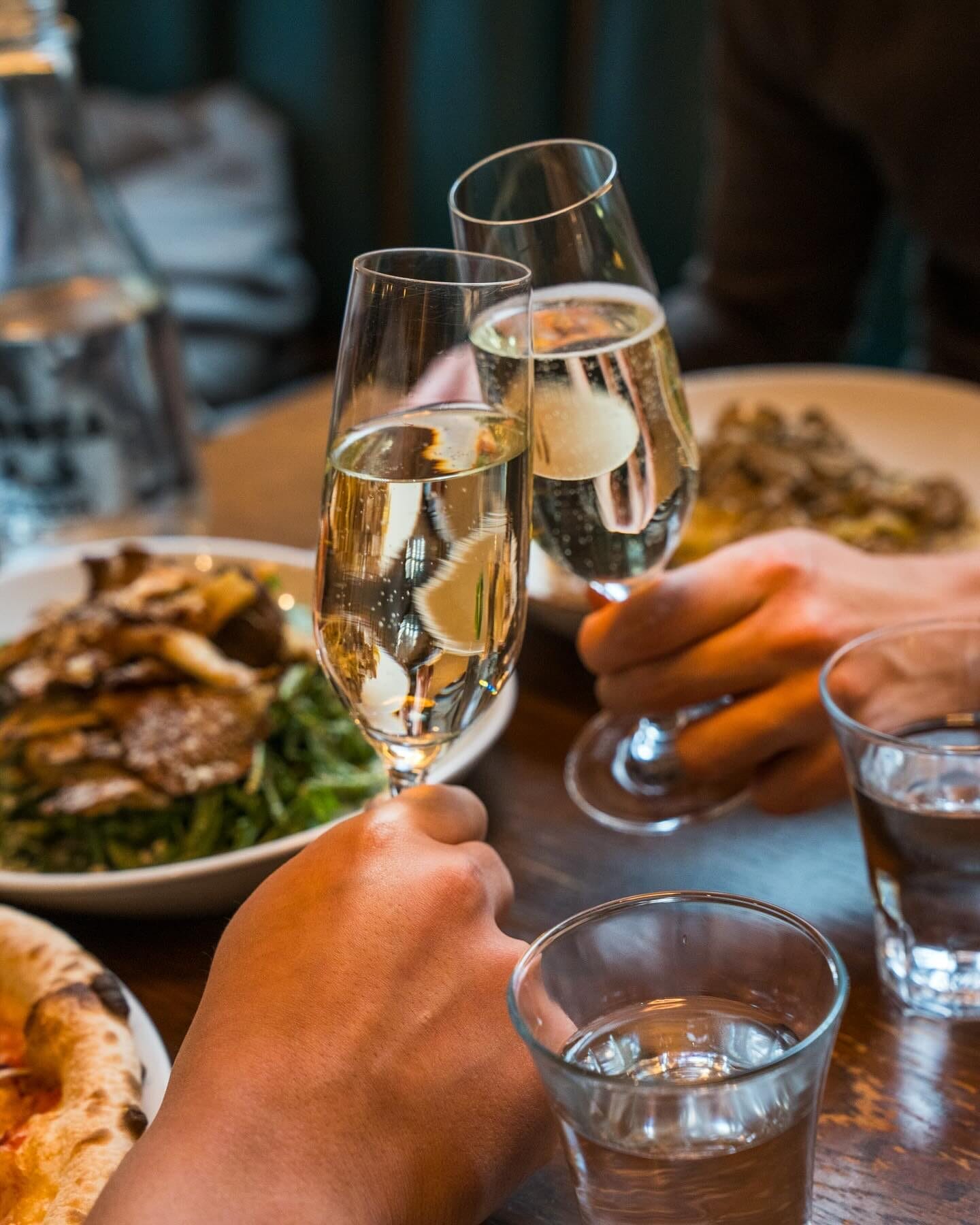 Two people clink champagne glasses over a wooden table set with plates of food, including a salad and a pizza. The warm lighting and close-up perspective capture a celebratory and intimate dining moment. | Open on Easter