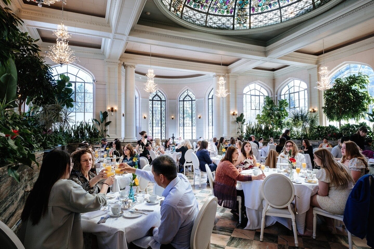 A bright, elegant dining hall with high ceilings, arched windows, and a stained-glass dome, filled with people enjoying a meal. Chandeliers hang from the ceiling, and lush greenery surrounds the space, creating a refined yet lively atmosphere.