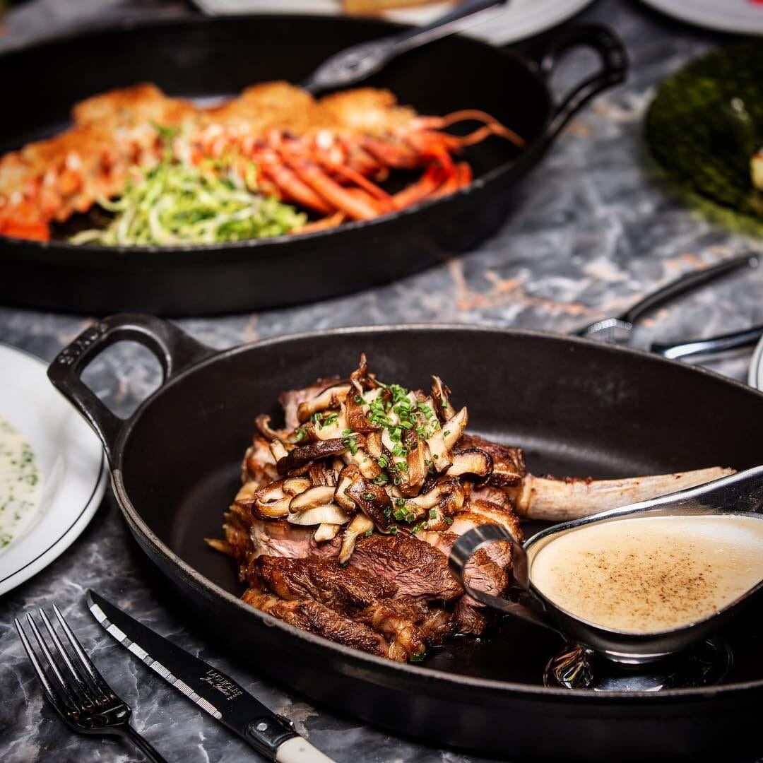 A cast-iron skillet holds a perfectly cooked tomahawk steak topped with sautéed mushrooms and garnished with chopped herbs, served alongside a rich, creamy sauce. In the background, a second skillet features a golden-crusted lobster tail over a bed of fresh greens. | Open on Easter