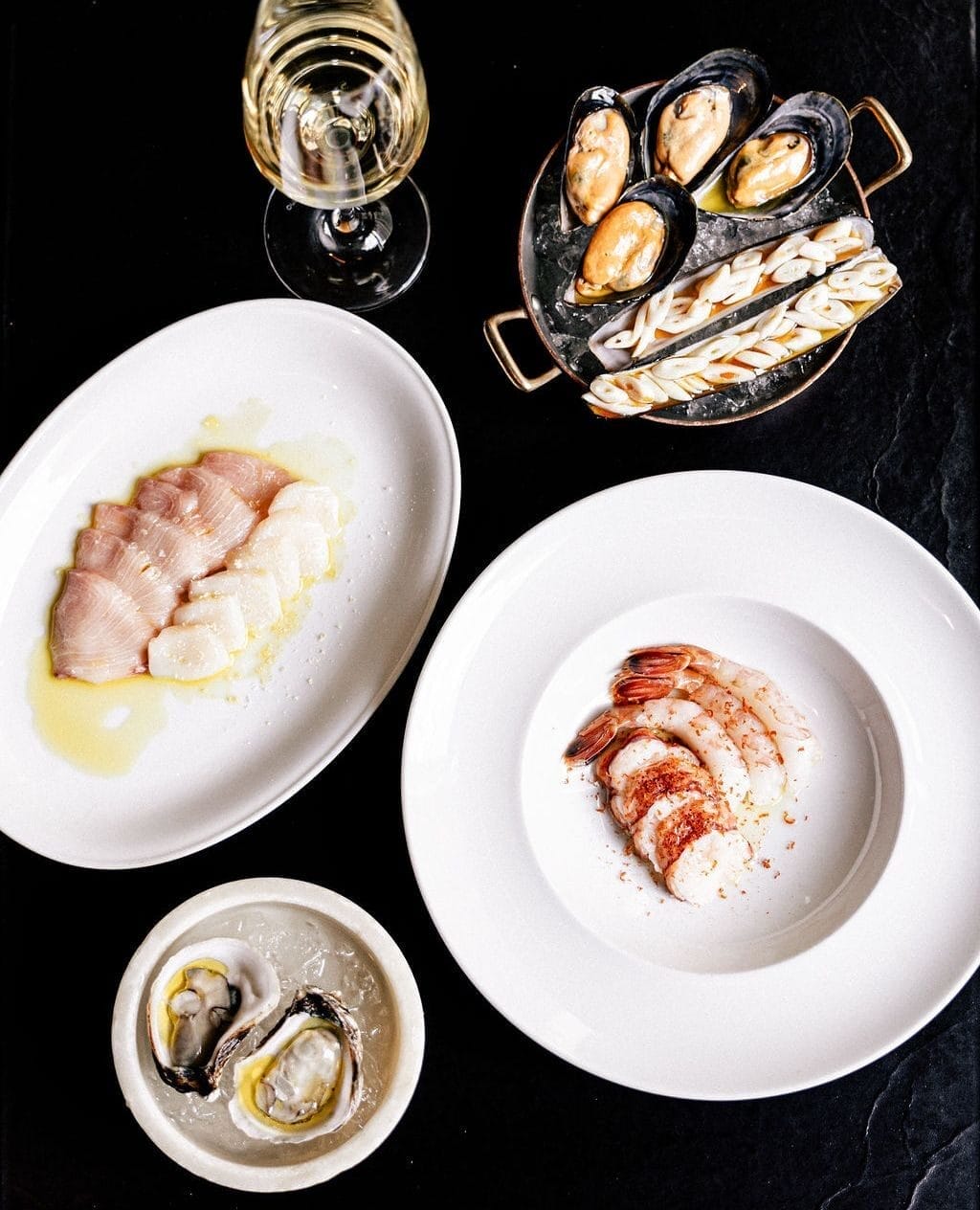 A top-down view of a seafood spread on a black table, featuring plates of fresh oysters on ice, thinly sliced raw fish drizzled with olive oil, shrimp and lobster pieces with seasoning, and a pan of mussels and razor clams. A glass of white wine is also placed beside the dishes.