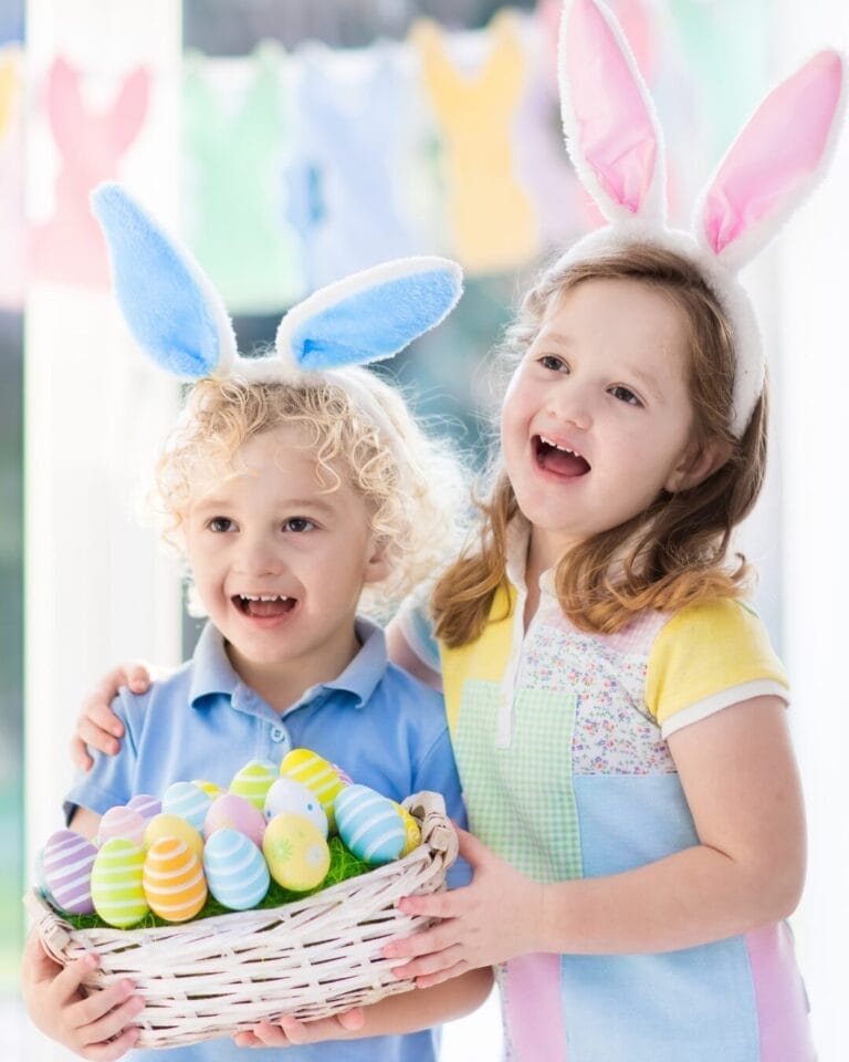 Two young children wearing bunny ears smile while holding a white wicker basket filled with colorful Easter eggs. The child on the left wears a blue shirt with matching blue bunny ears, while the child on the right wears a pastel-colored dress with pink bunny ears. A festive bunny-shaped garland hangs in the background, adding to the cheerful Easter setting.