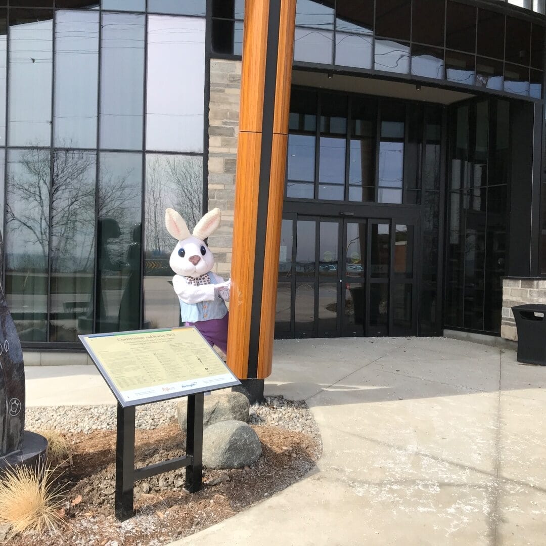 A person dressed in an Easter Bunny costume stands partially hidden behind a wooden pillar outside a modern glass building. The bunny is wearing a light blue vest with a patterned bow tie and purple pants. In the foreground, there is an informational sign mounted on a black stand, surrounded by rocks and dry grass landscaping. The background features large windows reflecting trees and an overcast sky.