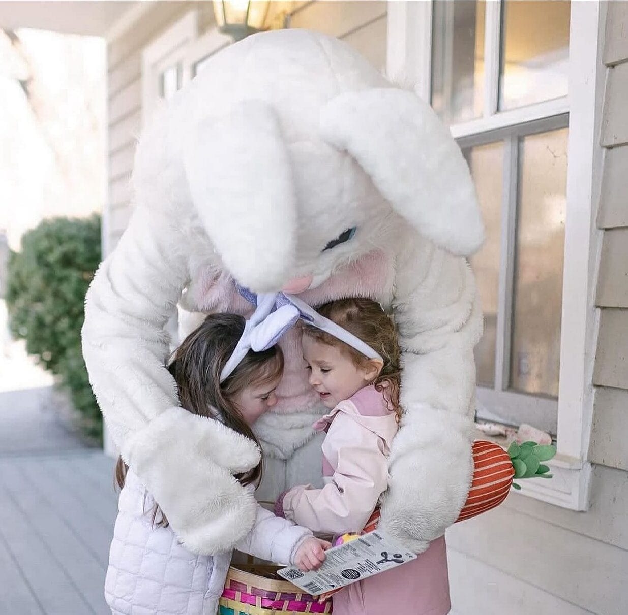 A person in an Easter Bunny costume embraces two young girls on a wooden porch outside a house. The girls, dressed in light-colored jackets, are smiling while looking at a colorful woven basket filled with Easter eggs and a flyer. One girl wears bunny ears, and the Easter Bunny holds a plush carrot prop. The background features a window, a lantern, and greenery.
